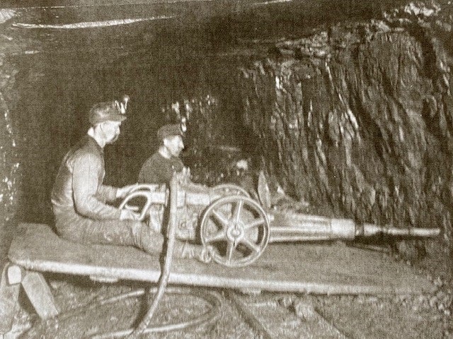 Historic photo of two miners straddling a punching machine in a coal mine.