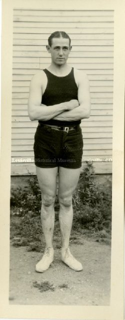Historic photo of man in tank top and shorts standing with arms crossed.
