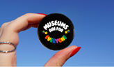 a woman's hand holds a black pin with rainbow words reading, 
